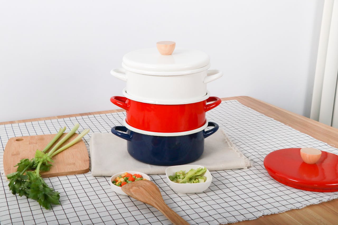 A stack of colorful cookware with ingredients and a wooden spoon on a kitchen table.